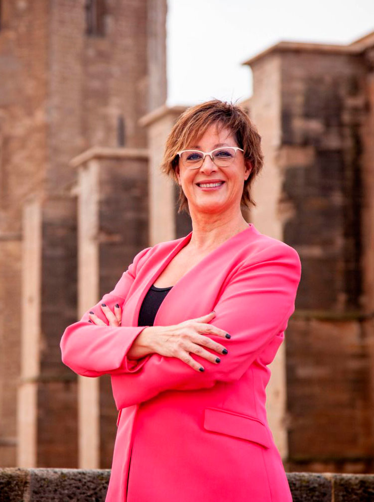 Una mujer con gafas y una americana rosa brillante está de pie al aire libre con los brazos cruzados, sonriendo con confianza. Al fondo, un edificio de piedra con altas columnas.